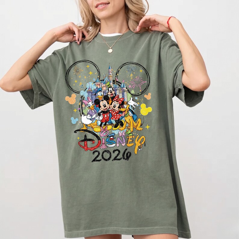 K&ouml;nnte beinhalten: Olivgr&uuml;nes T-Shirt mit einem farbenfrohen Disney-Motiv mit Mickey und Minnie Mouse, Donald Duck und anderen Charakteren. Die Grafik enth&auml;lt den Text "Disney 2026" und Feuerwerk.