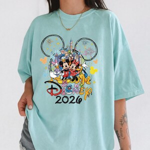 K&ouml;nnte beinhalten: Hellblaues T-Shirt mit einem farbenfrohen Disney-Motiv. Das Design zeigt Micky und Minnie Maus, Donald Duck und Goofy vor einem Schloss. Das Wort "Disney" und das Jahr "2026" sind ebenfalls auf dem T-Shirt aufgedruckt.