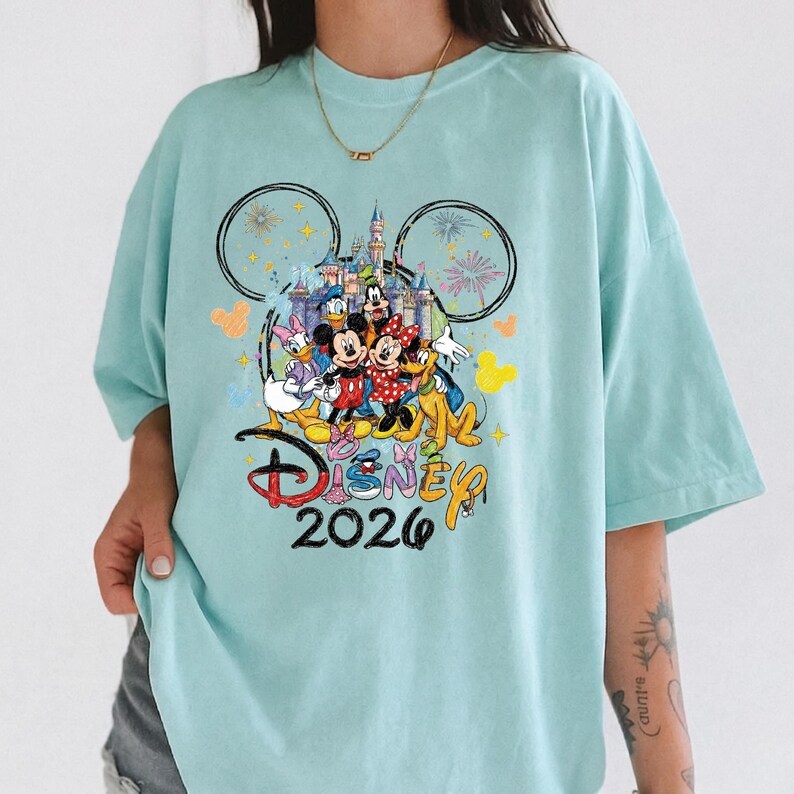 K&ouml;nnte beinhalten: Hellblaues T-Shirt mit einem farbenfrohen Disney-Motiv. Das Design zeigt Micky und Minnie Maus, Donald Duck und Goofy vor einem Schloss. Das Wort "Disney" und das Jahr "2026" sind ebenfalls auf dem T-Shirt aufgedruckt.