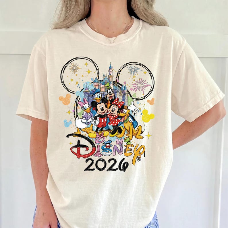 K&ouml;nnte beinhalten: Cremefarbenes T-Shirt mit einem farbenfrohen Disney-Motiv. Das Design zeigt Micky und Minnie Maus, Donald Duck, Daisy Duck, Goofy und ein Schloss. Der Text "Disney 2026" ist ebenfalls zu sehen.