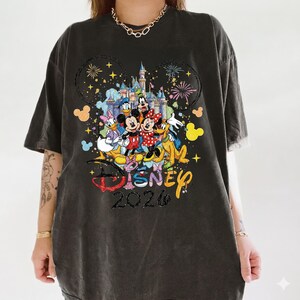 K&ouml;nnte beinhalten: Dunkelgraues T-Shirt mit einem farbenfrohen Disney-Motiv. Das Design zeigt Micky und Minnie Maus, Donald Duck und andere Charaktere vor einem Schloss. Das Wort "Disney" und das Jahr "2026" sind ebenfalls zu sehen.