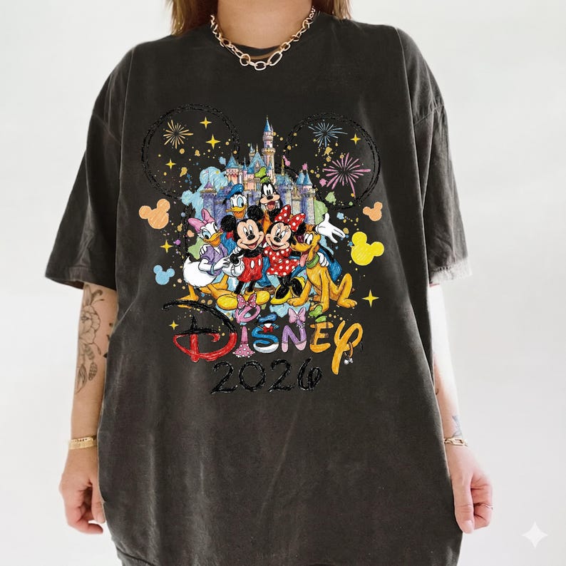 K&ouml;nnte beinhalten: Dunkelgraues T-Shirt mit einem farbenfrohen Disney-Motiv. Das Design zeigt Micky und Minnie Maus, Donald Duck und andere Charaktere vor einem Schloss. Das Wort "Disney" und das Jahr "2026" sind ebenfalls zu sehen.