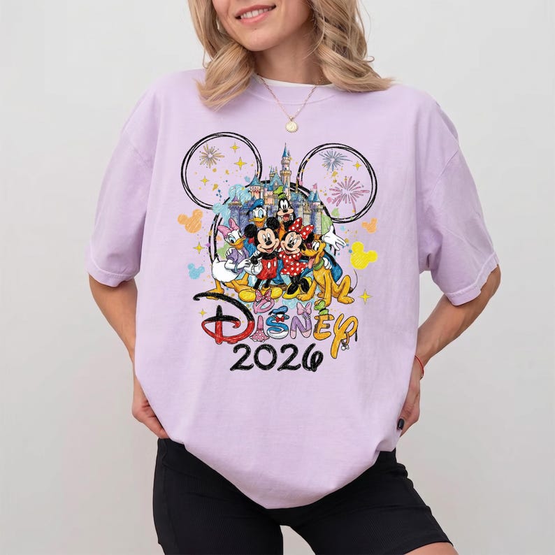 K&ouml;nnte beinhalten: Helllila T-Shirt mit einem farbenfrohen Disney-Motiv. Das Motiv zeigt Micky und Minnie Maus, Donald Duck und Goofy, mit dem Text "Disney 2026" unter den Figuren.