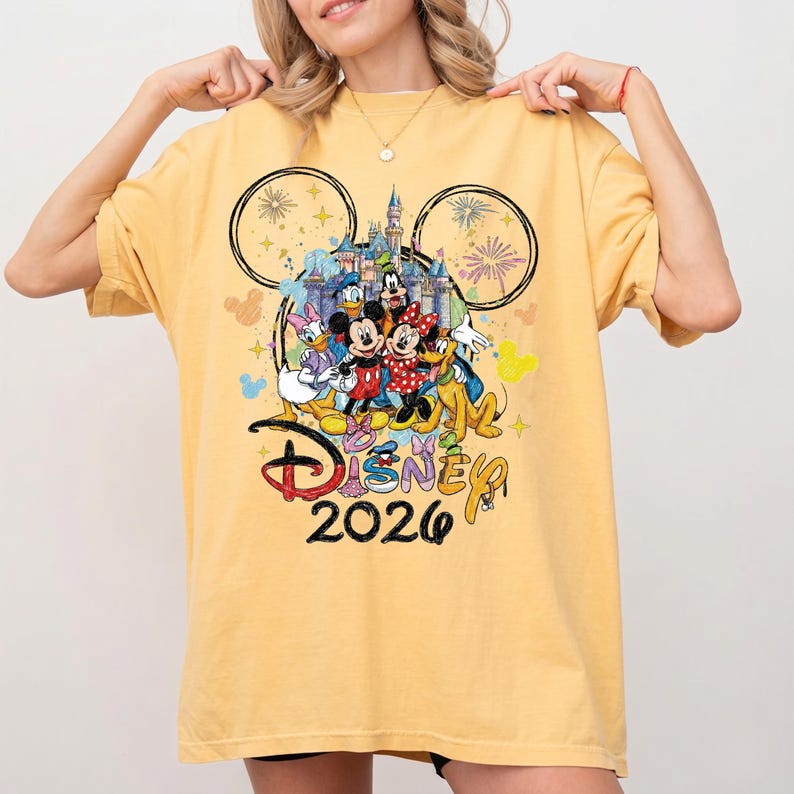 K&ouml;nnte beinhalten: Senfgelbes T-Shirt mit einem farbenfrohen Aufdruck von Disney-Figuren, darunter Mickey und Minnie Mouse, Donald Duck und Goofy. Das Design zeigt ein Schloss, Feuerwerk und den Text "Disney 2026".
