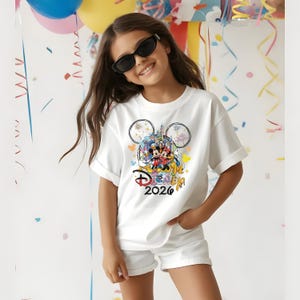 K&ouml;nnte beinhalten: Wei&szlig;es T-Shirt mit einem farbenfrohen Disney-Motiv mit Mickey und Minnie Mouse, dem Disney-Schloss und dem Text "Disney 2026". Das Shirt hat hochgekrempelte &Auml;rmel und wird mit wei&szlig;en Shorts getragen. Die Person im Bild tr&auml;gt eine schwarze Sonnenbrille.