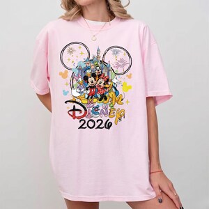 K&ouml;nnte beinhalten: Hellrosa T-Shirt mit einem farbenfrohen Disney-Motiv. Das Design zeigt Micky und Minnie Maus, Donald Duck und Goofy, mit einem Schloss und Feuerwerk. Das Wort "Disney" und das Jahr "2026" sind ebenfalls abgebildet.