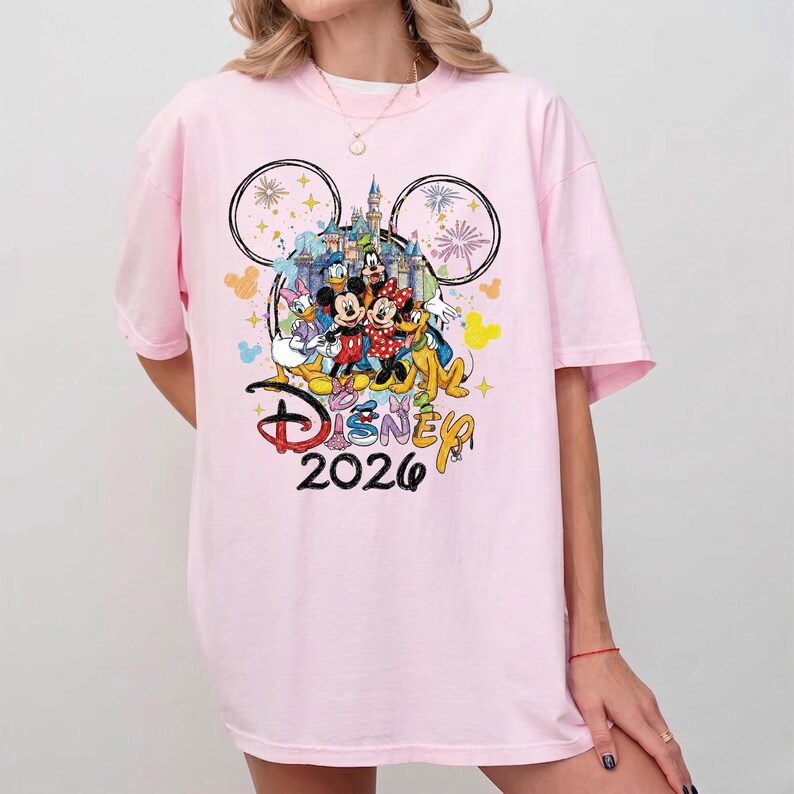 K&ouml;nnte beinhalten: Hellrosa T-Shirt mit einem farbenfrohen Disney-Motiv. Das Design zeigt Micky und Minnie Maus, Donald Duck und Goofy, mit einem Schloss und Feuerwerk. Das Wort "Disney" und das Jahr "2026" sind ebenfalls abgebildet.