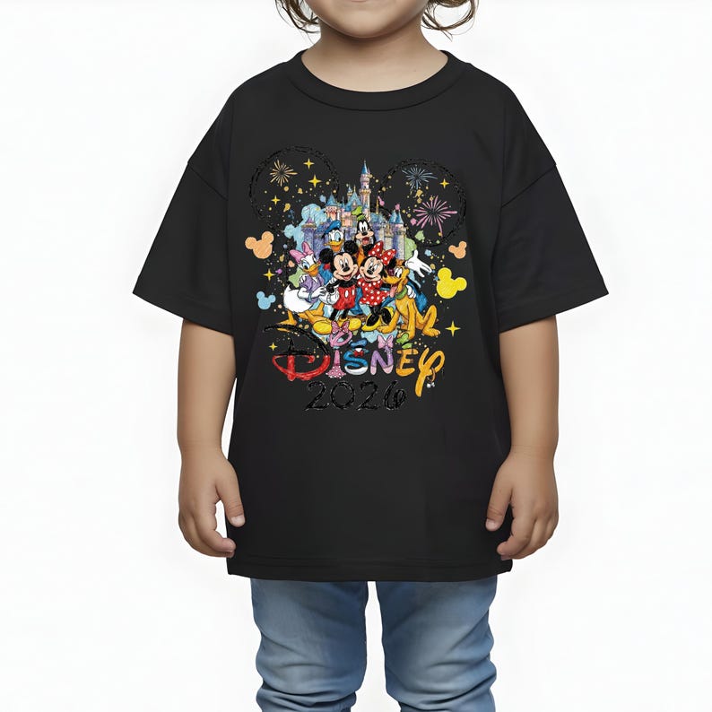 K&ouml;nnte beinhalten: Schwarzes T-Shirt mit einer bunten Grafik von Disney-Figuren, darunter Micky und Minnie Maus, vor einem Schloss. Der Text "Disney 2026" ist ebenfalls auf dem T-Shirt abgebildet. Das T-Shirt wird von einem Kind getragen.