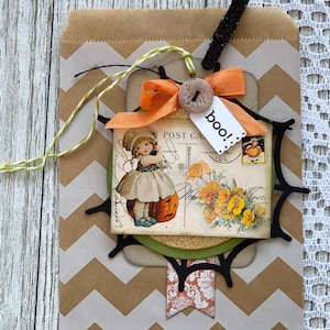 Puede incluir: Una bolsa de papel marrón con un patrón de chevron blanco y marrón. Una telaraña negra con un círculo verde y dorado en el centro está unida a la bolsa. Una postal vintage con una niña con un vestido blanco y una calabaza está unida a la telaraña. Una cinta negra y blanca con un botón blanco y un lazo naranja está unida a la telaraña. Una etiqueta blanca con la palabra "boo!" está unida a la cinta.