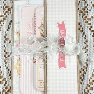 May include: A pink and white gingham card with a pink ribbon and a white lace bow. The card has a pink banner with the text "gingerbread" on it. The card is decorated with a gingerbread man and a Christmas tree.