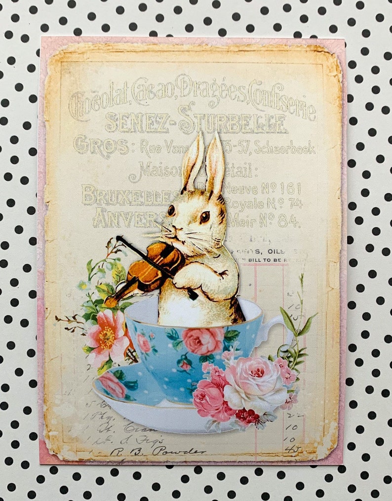 May include: A vintage illustration of a white rabbit playing a violin while sitting in a blue and white teacup with pink roses. The image is on a pink and cream background with text that reads "Chocolat Cacao, Drag&eacute;es Confiserie, Seneze-Sturbeelge, Gros: Rue Van 15-57, Scherbeck, Maison Retail: Neuve No 161, Royale No 74, Meir No 84."