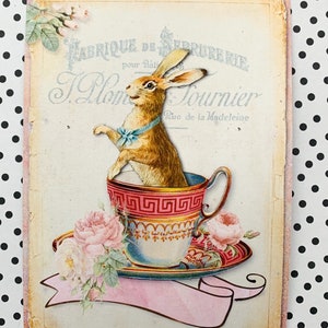 May include: A vintage illustration of a brown rabbit sitting in a teacup with a blue bow tie. The teacup is on a saucer with pink roses and green leaves. The background is a cream-colored paper with the text "Fabrique de Serrurerie pour B&acirc;timents J. Plom  Tournier Rue de la Madeleine" in black and blue lettering.
