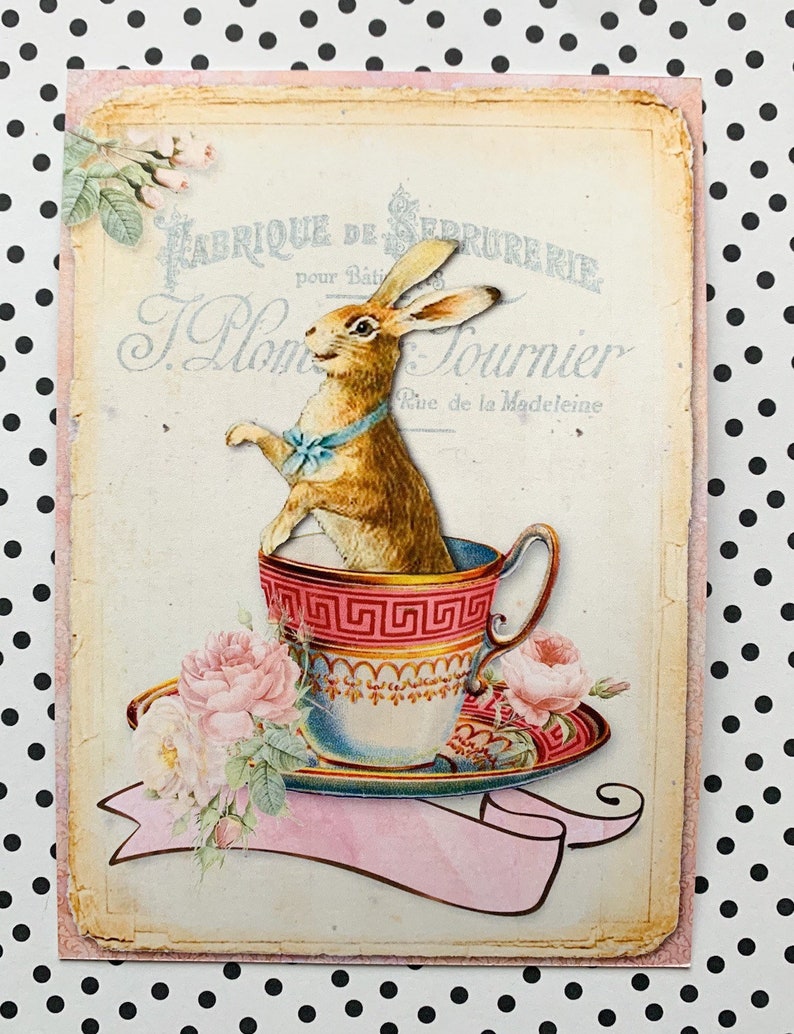 May include: A vintage illustration of a brown rabbit sitting in a teacup with a blue bow tie. The teacup is on a saucer with pink roses and green leaves. The background is a cream-colored paper with the text "Fabrique de Serrurerie pour B&acirc;timents J. Plom  Tournier Rue de la Madeleine" in black and blue lettering.