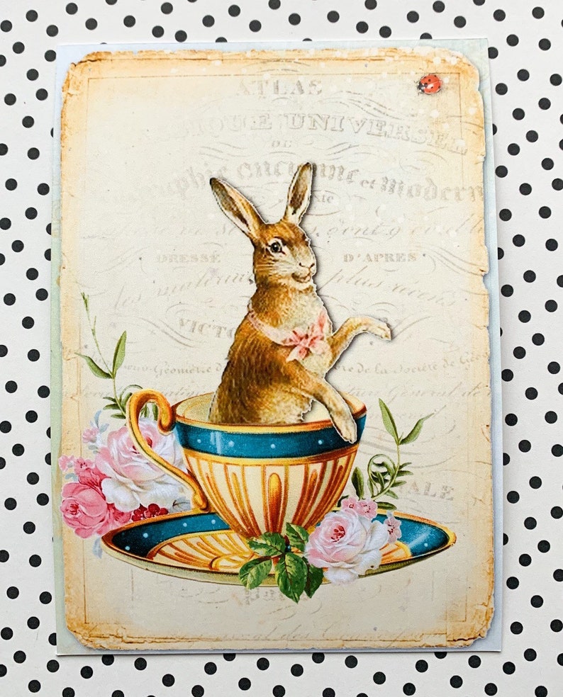 May include: A vintage illustration of a brown rabbit sitting in a blue and gold teacup. The rabbit is wearing a pink bow and the teacup is surrounded by pink and white flowers.