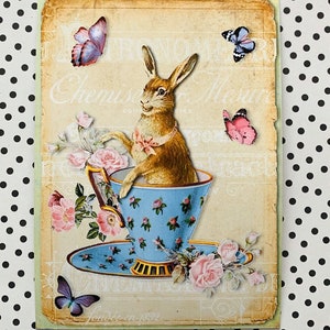 May include: A vintage illustration of a brown rabbit sitting in a blue teacup with a saucer. The teacup and saucer are decorated with pink roses. There are pink, blue, and purple butterflies flying around the rabbit.