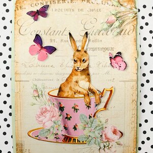 May include: A vintage-style illustration of a brown rabbit sitting in a pink teacup with a saucer. The teacup is surrounded by pink roses and green leaves. The background is a faded, cream-colored paper with French text.