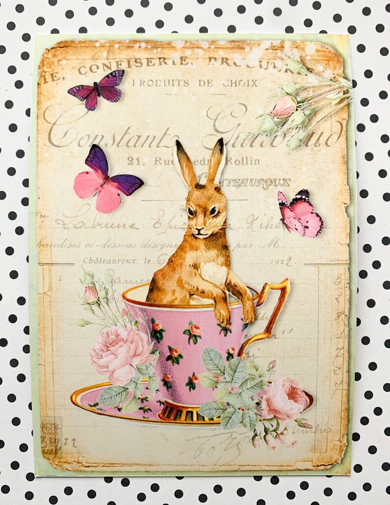 May include: A vintage-style illustration of a brown rabbit sitting in a pink teacup with a saucer. The teacup is surrounded by pink roses and green leaves. The background is a faded, cream-colored paper with French text.
