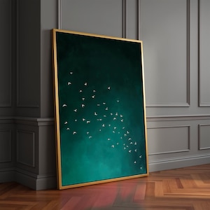 May include: A framed art print featuring a flock of white birds against a dark teal and green gradient background. The artwork is encased in a gold-colored frame, leaning against a gray wall and wooden floor.