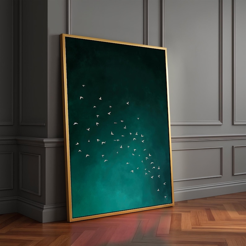 May include: A framed art print featuring a flock of white birds against a dark teal and green gradient background. The artwork is encased in a gold-colored frame, leaning against a gray wall and wooden floor.