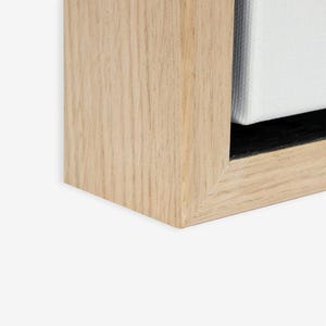 May include: Close-up of a corner of a light-colored wooden picture frame. The frame has a natural wood grain finish and a black interior. A white canvas is partially visible within the frame.