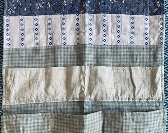 Op de afbeelding: Een stoffen organizer met meerdere vakken. Het bovenste gedeelte is wit, gevolgd door een blauw patroon met dromenvangers en veren. Daaronder strepen in wit en blauw, dan een ruitpatroon en tot slot vakken.