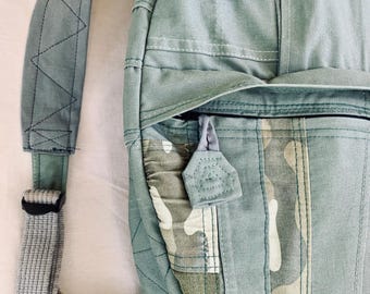 May include: A close-up of a light green backpack with a camouflage pattern. The backpack has a button closure, a small pocket, and adjustable straps. The stitching is visible throughout the design. The backpack is made of a durable fabric.