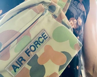 May include: A camouflage fanny pack with the words "AIR FORCE" on a rectangular patch. The bag features a zipper closure and adjustable strap. The pattern includes green, brown, and tan circular shapes.