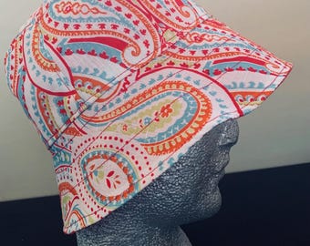 May include: A paisley print bucket hat in red, orange, blue, and yellow on a white background. The hat is displayed on a gray mannequin head, showcasing the hat's shape and design. The hat's brim is slightly angled.