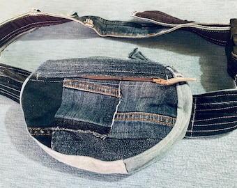May include: A denim fanny pack with a patchwork design. The bag features a zippered main compartment and an adjustable strap with a black buckle. The denim is a mix of light and dark blue washes, with brown stitching and a light-colored zipper pull.