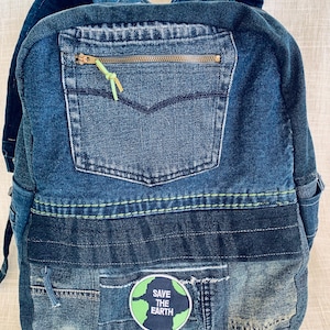 May include: A denim backpack in various shades of blue. The front pocket features a zip with a green pull. A patch on the front reads "Save the Earth." The backpack is made from recycled denim.