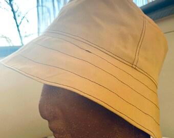 May include: Tan bucket hat with a wide brim and black stitching details. The hat has a structured design and a classic, casual style. The hat is made of a durable fabric, suitable for sun protection and outdoor activities.