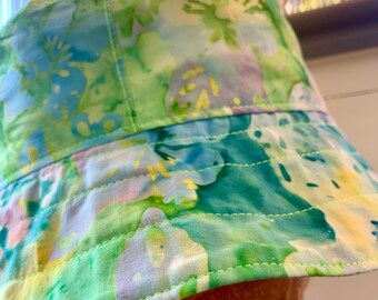 May include: A colorful bucket hat with a floral pattern in shades of green, blue, yellow, and purple. The hat has a wide brim and is made of a soft fabric. The stitching is visible around the brim and the crown of the hat.