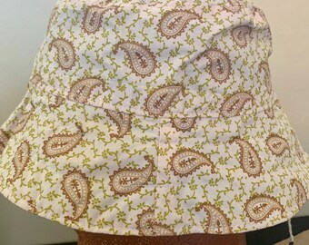 May include: A white bucket hat with a paisley pattern in brown and green. The hat has a wide brim and is made of a soft fabric. The paisley design is repeated across the entire hat.