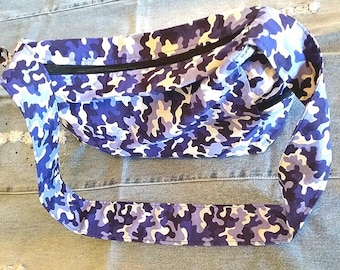 May include: A crossbody bag featuring a blue, purple, white, and light gray camouflage pattern. The bag has a wide, integrated shoulder strap and a black zipper closure running along the top edge of the main compartment.