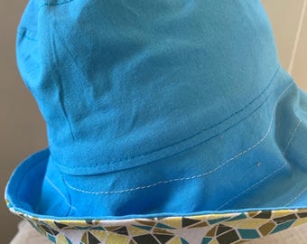 May include: A blue bucket hat with a patterned brim. The hat is a solid light blue color with white stitching. The brim features a geometric pattern in yellow, teal, and brown. The hat is made of a soft fabric.