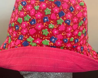 May include: A bright pink bucket hat with a floral pattern in pink, blue, and green. The brim is a solid pink color. The hat is displayed on a mannequin head.