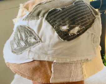 May include: A patchwork bucket hat in neutral tones. The hat features various fabric patches in beige, tan, olive green, and cream, with visible stitching and frayed edges. The brim is a solid tan color. This hat is a unique, handcrafted accessory.