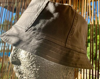 May include: A gray bucket hat with a structured design. The hat features a wide brim and is made of a durable fabric. White stitching accents the seams. The hat is displayed on a stone head.