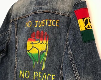 May include: A blue denim jacket with a raised fist painted on the back. The fist is painted in red, yellow, and green. The words "NO JUSTICE NO PEACE" are written in yellow on the back of the jacket. A red, yellow, and green patch with a peace sign is sewn on the right sleeve.