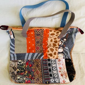 Puede incluir: Un bolso tote patchwork con una variedad de patrones y colores de tela, incluyendo naranja, marrón y azul. El bolso tiene una correa azul y una correa marrón, y un cierre de cremallera.