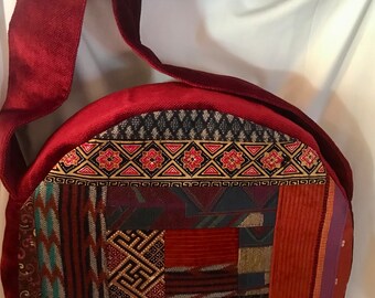 May include: A round, red, patchwork fabric bag with a long, red strap. The bag is made of different fabrics with geometric patterns in shades of red, brown, blue, and green.