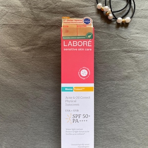 May include: A pink and white LABORE sensitive skin care box with the text "Acne & Oil Correct Physical Sunscreen SPF 50+ PA++++". A black hair tie with white pearl beads is next to the box.