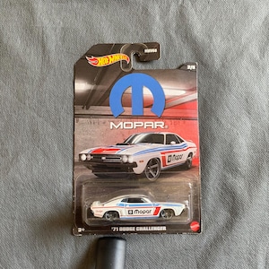 May include: A Hot Wheels die-cast toy car of a white and blue '71 Dodge Challenger with Mopar branding. The packaging features the Hot Wheels logo, Mopar logo, and a graphic of the car. The car is approximately 3 inches long.