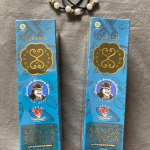 以下が含まれることがあります： 金色のエンブレムとテキストが付いた、Sanga Sangaハーブオイルの2つの青い箱。箱には「Classic」と書かれており、男性の肖像画が描かれています。白と金のビーズが付いた黒いヘアタイが一番上にあります。