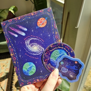 May include: A rectangular greeting card and two circular stickers featuring space themes. The dark blue designs include a spiral galaxy, planets, comets, and stars with the text "beautiful things are out there, go out and find them" printed.