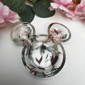 May include: A clear glass trinket dish shaped like Mickey Mouse ears, filled with pressed flowers and leaves in shades of green, red, and white.