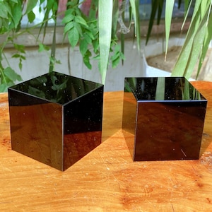 Op de afbeelding: Twee zwarte, glanzende, kubusvormige objecten staan op een houten oppervlak. De kubussen reflecteren licht, waardoor een spiegelachtig effect ontstaat. De kubussen staan naast elkaar, met een natuurlijke achtergrond van groen gebladerte.