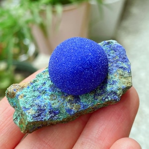 Op de afbeelding: Een helderblauwe minerale bol rust op een getextureerde, veelkleurige rotsformatie. De rots toont tinten groen, blauw en bruin. Het oppervlak van de bol heeft een korrelige textuur.