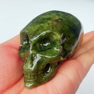 May include: A carved green skull figurine. The skull is a vibrant green color with darker green marbling throughout. The skull's features, including eye sockets, nose cavity, and teeth, are clearly defined. The figurine is held in a hand.