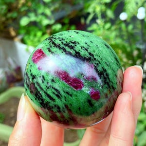 May include: A polished sphere of green zoisite with ruby inclusions. The gemstone features a vibrant green base with black streaks and patches of deep red ruby crystals. The sphere is held in a hand against a blurred green background.